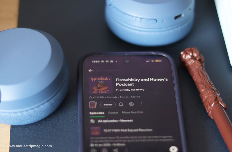 A phone playing a Harry Potter podcast lay on a desk beside some blue headphones and a plastic wand.